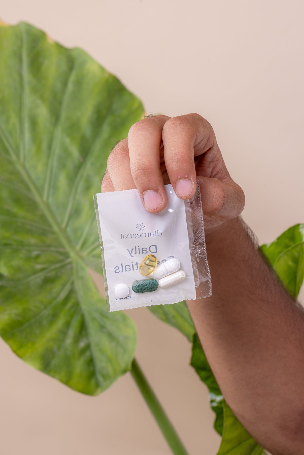 A hand holding a small clear vitameenat daily essentials sachet containing 5 supplements, with a large green tropical leaf and a beige background.
