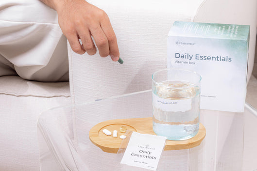 Man sitting on a couch with a glass of water, a box of VItameenat 'Daily Essentials' on a small table.